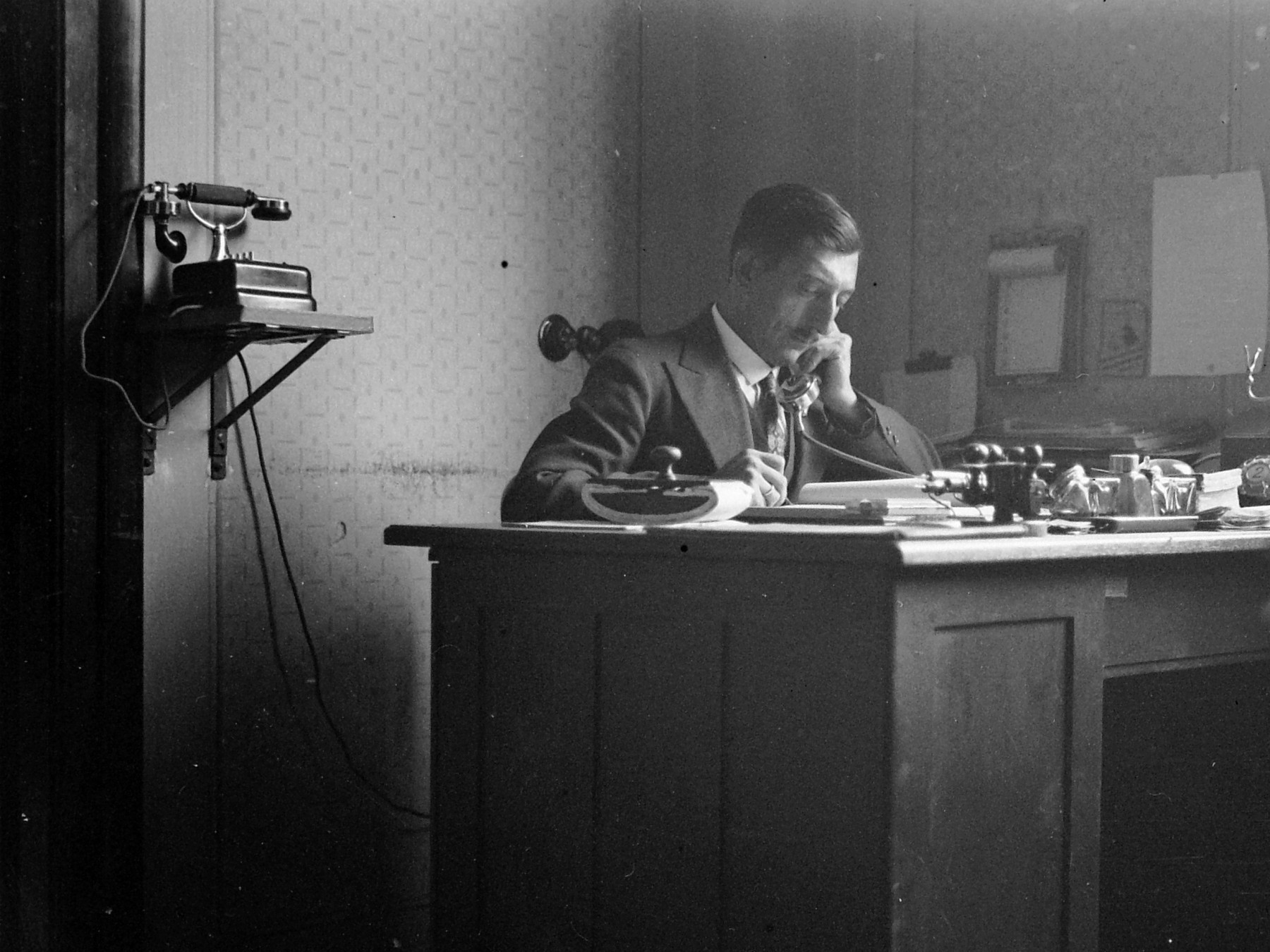 Чёрно-белая фотография мужчины за письменным столом с телефоном и канцелярскими принадлежностями.